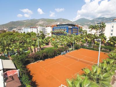 Kort tenisowy z ceglanym podłożem otoczony palmami, w tle budynek hotelu.