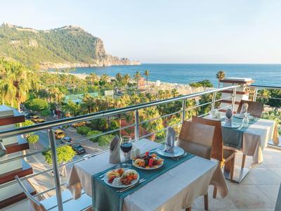 Balkon mit eingedeckten Tischen, Blick auf Meer, Palmen und grüne Hügel im Hintergrund.