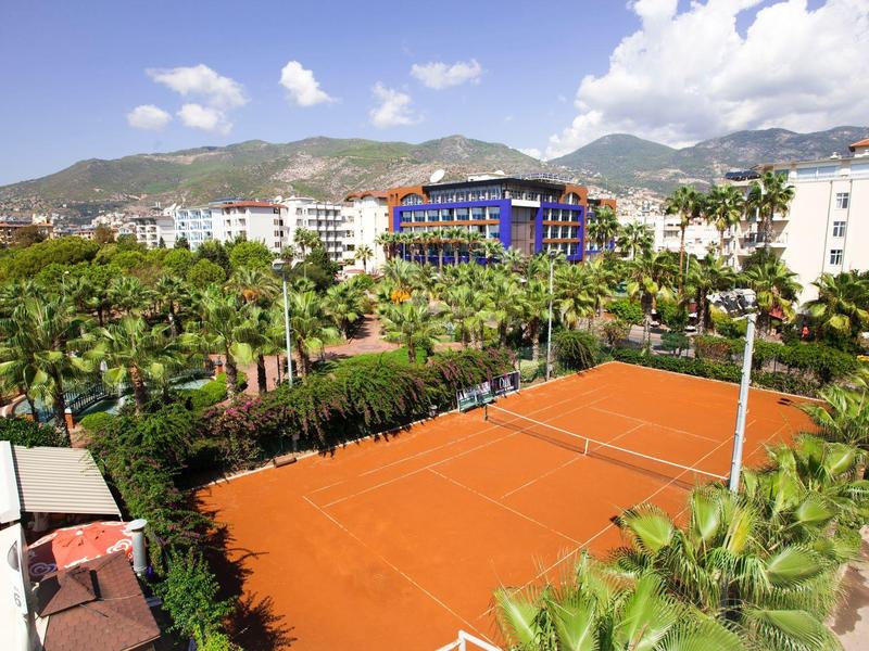 Rotes Tennisfeld umgeben von Palmen und Gebäuden vor bewölktem Bergpanorama.