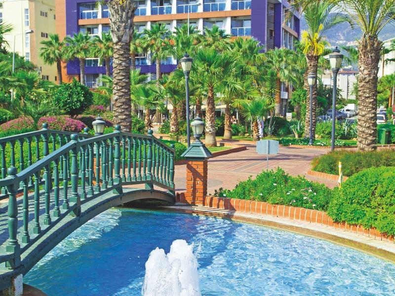 Ein blauer Hotelbau mit Palmen davor, einem kleinen Bogenbrücke und einem Springbrunnen im Pool.