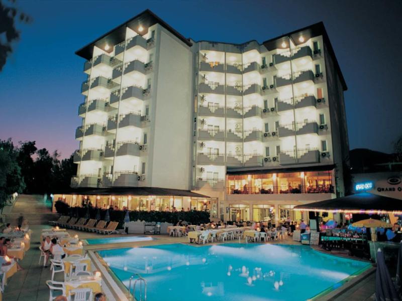 Hotel mit beleuchtetem Außenpool, vielen Gästen und weißem sechsstöckigem Gebäude bei Abenddämmerung.