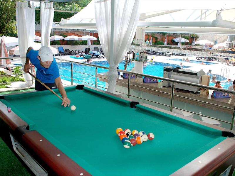 Person spielt Billard an einem großen grünen Tisch neben einem Schwimmbad mit Sonnenschirmen.