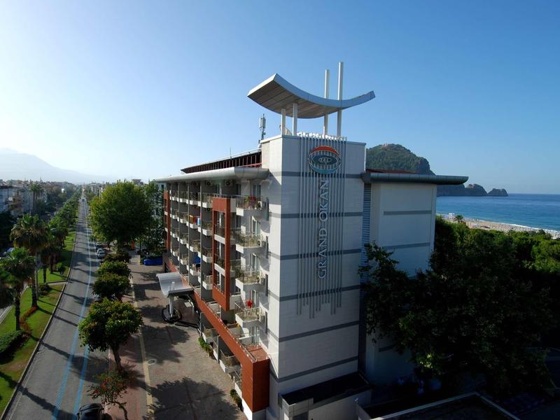 Langes Hotelgebäude an Strandpromenade mit Palmen, blauem Himmel und Meer im Hintergrund.