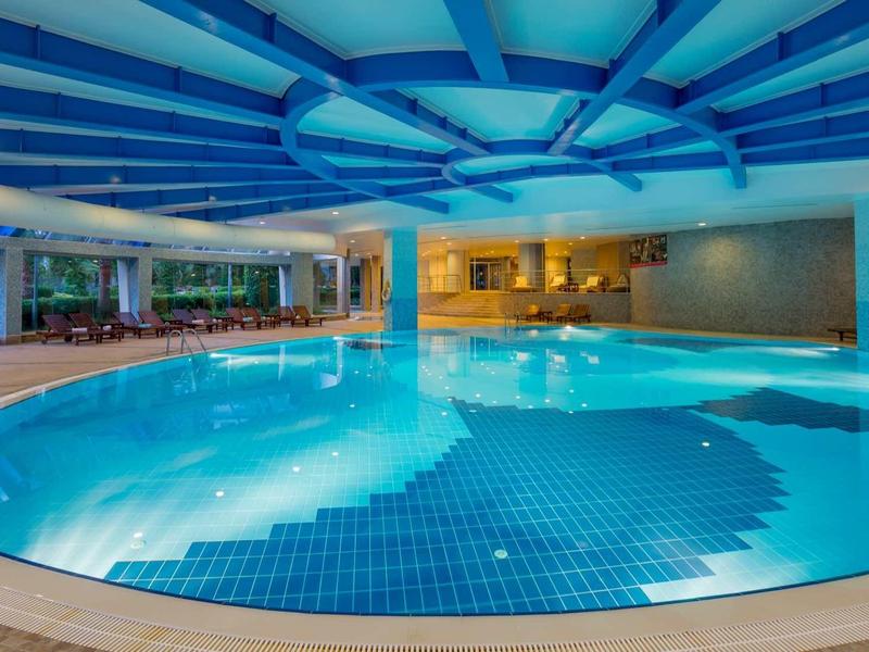 Velký krytý bazén s modrou vodou a bílými lehátky u okraje.