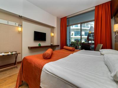 Modernes Hotelzimmer mit großem Bett, orangefarbenen Vorhängen und Balkon mit Stadtblick.