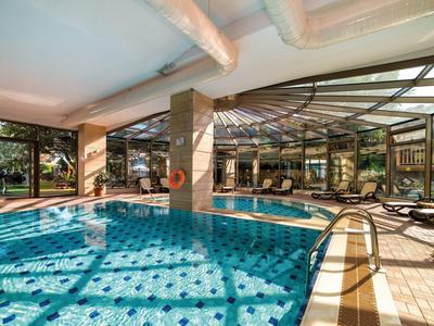Helles Hallenbad mit blauem Wasser, Glaswänden, Liegestühlen und heller Decke mit Rohren.