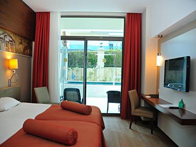 Helles Hotelzimmer mit zwei Betten, roten Vorhängen, Balkon und Fernseher an der Wand.