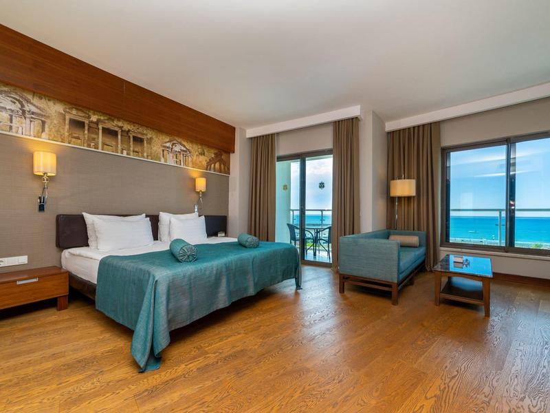 Hotelkamer met tweepersoonsbed, teal deken, fauteuil, balkon en uitzicht op zee.