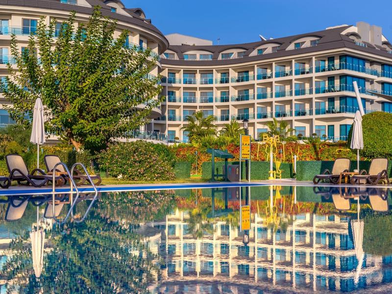 Hotelgebouw weerspiegeld in het rustige buitenzwembad met ligstoelen en groen.