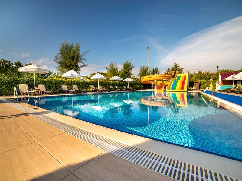 Buitenzwembad met glijbanen, omgeven door parasols en groen onder een zonnige hemel.