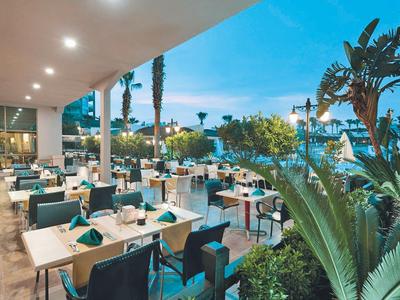 Área de restaurante de hotel al aire libre con mesas y sillas junto a plantas exuberantes al atardecer.