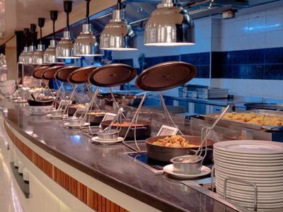 Buffet moderno con luces colgantes, múltiples platos calientes y pilas de platos en un hotel.