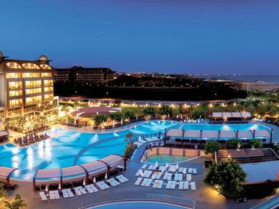 Großer beleuchteter Hotelpool mit Liegestühlen und Blick auf Meer und Gebäude bei Nacht.