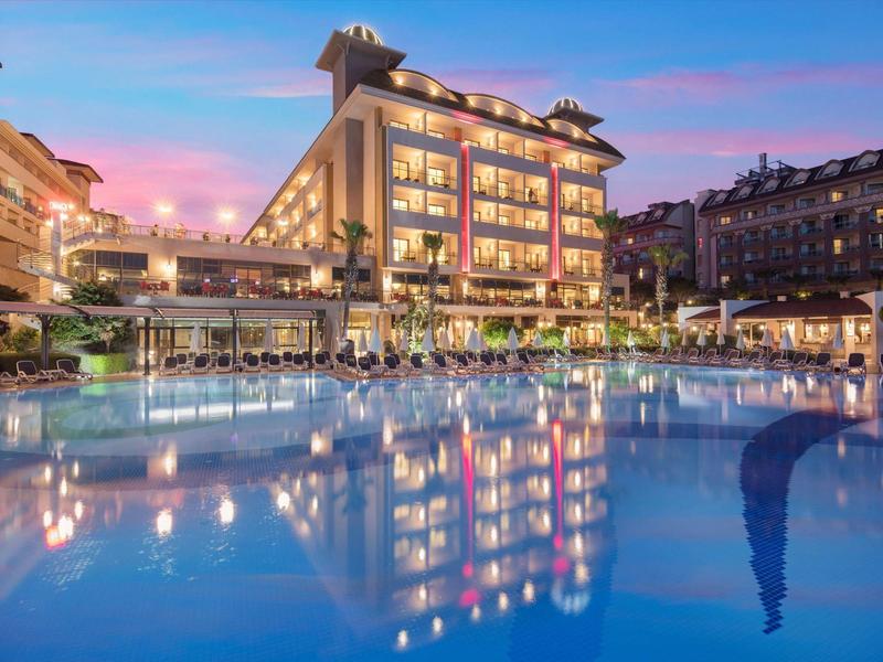 Vista nocturna de un hotel con piscina iluminada y reflejos en el agua.
