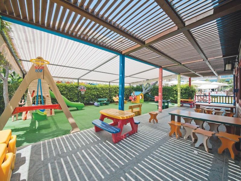 Área cubierta con bancos, mesas y un parque infantil con tobogán en el jardín.