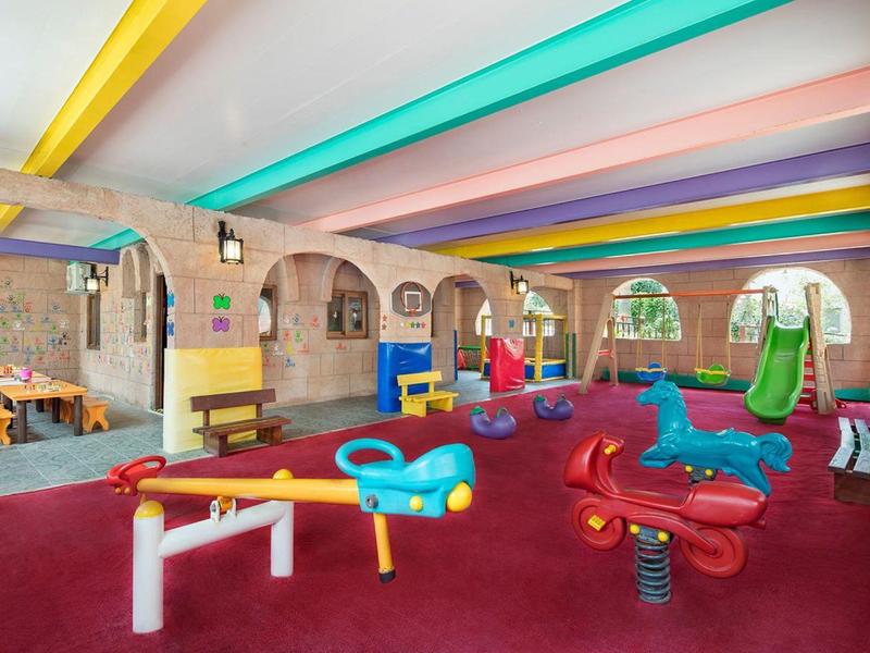Zona de juegos interior colorida con toboganes, balancines y vigas de techo multicolor en área de hotel.