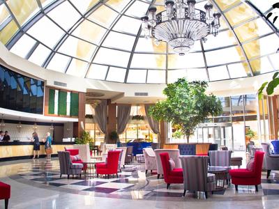 Recepción de hotel luminosa con techo de cristal, candelabro, árboles y sillas rojas.