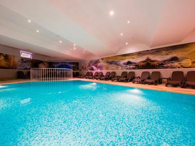 Hallenbad mit leuchtend blauem Wasser, Relaxliegen und Landschaftswandmalerei im Hintergrund.