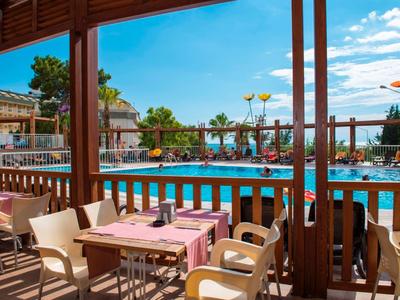 Terrasse mit Tischen und Stühlen neben einem Pool in einer Hotelanlage bei sonnigem Wetter.