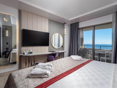 Camera d'hotel moderna con vista mare, letto matrimoniale e balcone con sedie.