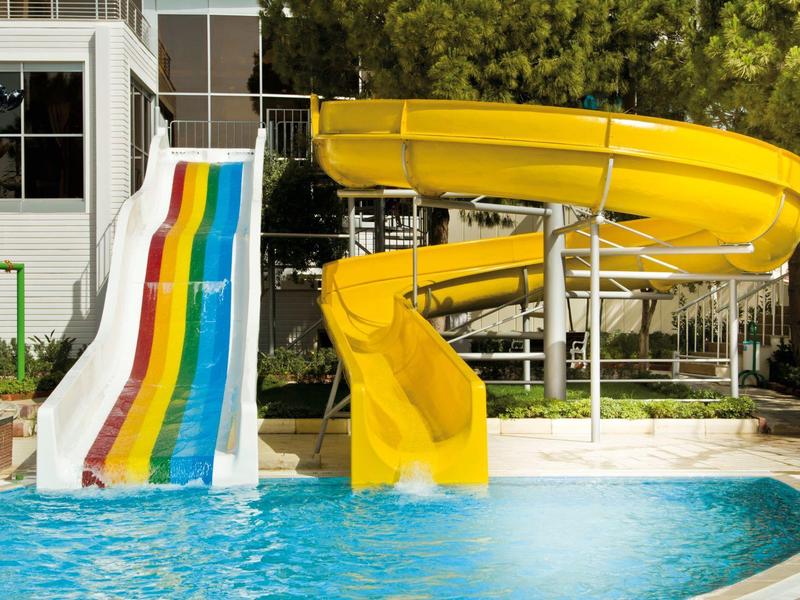 Dos toboganes de agua, uno amarillo en espiral y uno colorido recto, terminan en una piscina junto a un hotel.