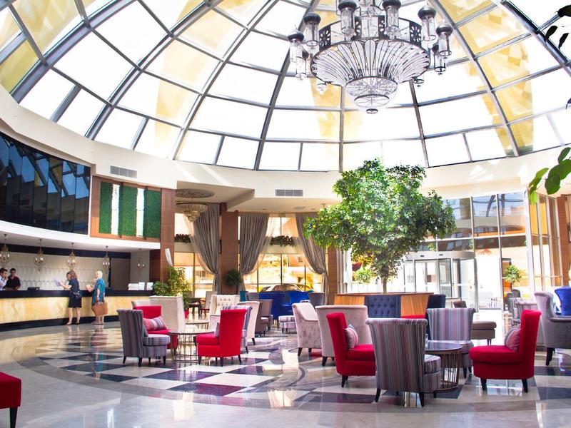 Recepción de hotel luminosa con techo de cristal, candelabro, árboles y sillas rojas.