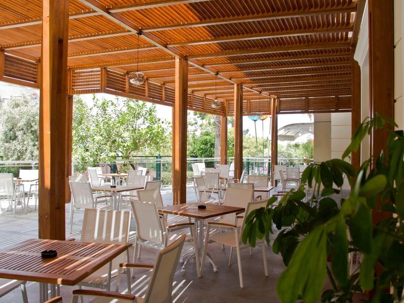 Terraza cubierta con techo de madera y mesas y sillas blancas, rodeada de plantas verdes.