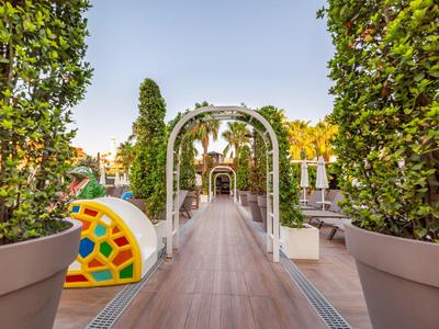 Ścieżka z łukami i dużymi roślinami w donicach, kolorowa rzeźba po lewej, na zewnątrz pod czystym niebem.