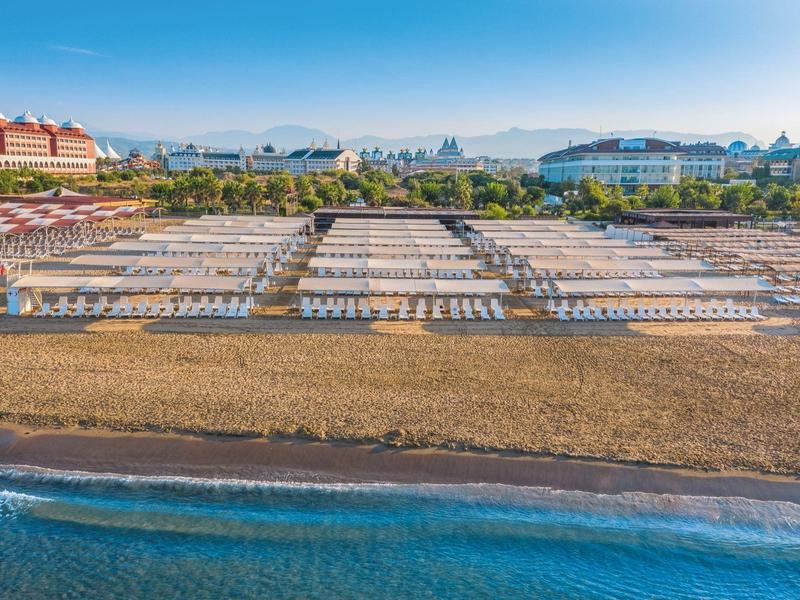 Plaża z rzędami białych leżaków przed hotelem i górami w tle.