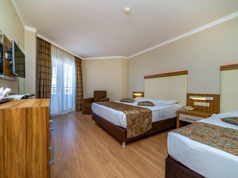 Jasny pokój hotelowy z dwoma łóżkami, drewnianą podłogą, fotelem i balkonem z zasłonami.