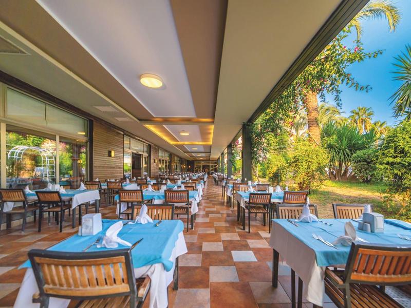 Restauracja na zewnątrz z nakrytymi stołami i drewnianymi krzesłami obok ogrodu w słoneczny dzień.