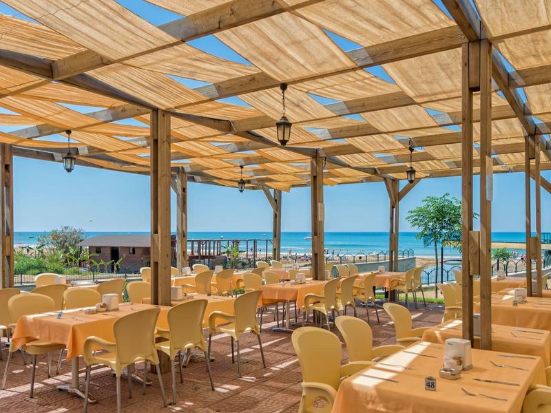 Salle à manger ouverte avec vue sur la plage et la mer, plafond en bois et nombreuses tables.