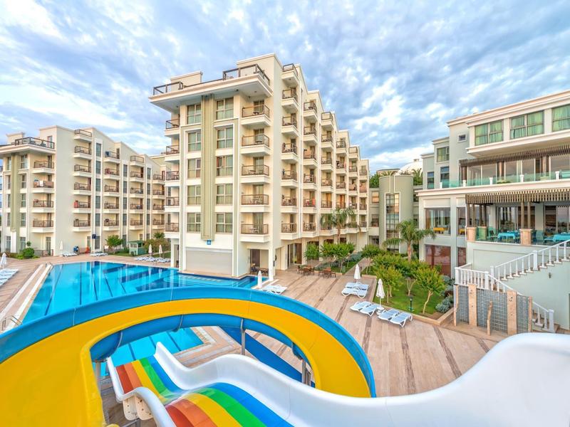 Complexe hôtelier moderne à ailes multiples avec grande piscine, toboggan et terrasse sous un ciel nuageux.