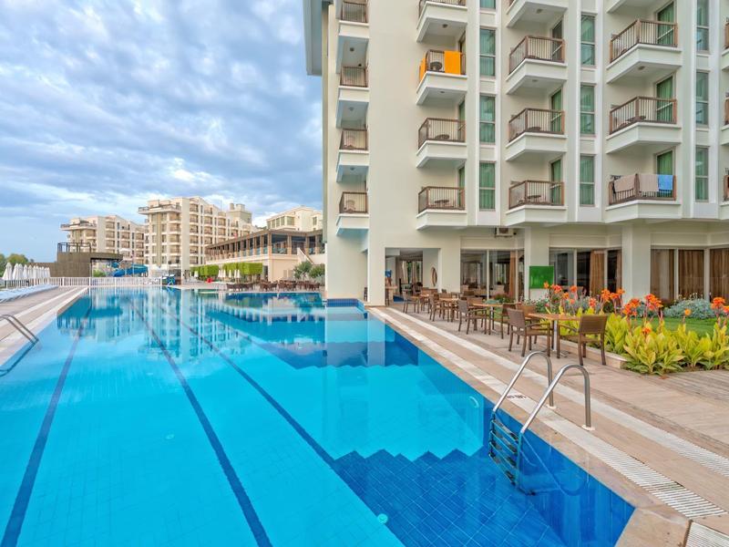 Grande piscine d'hôtel avec eau bleu clair à côté d'un bâtiment hôtelier à plusieurs étages sous un ciel nuageux.