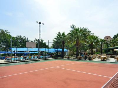 Kort tenisowy z czerwonym podłożem otoczony drzewami i budynkami w słoneczną pogodę.