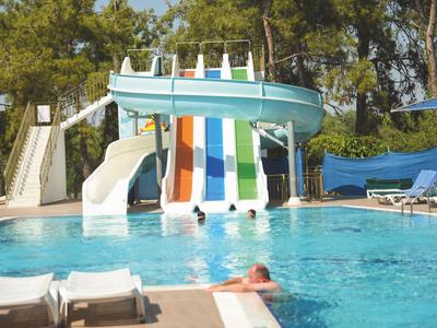 Schwimmbad mit bunten Wasserrutschen, blauem Wasser und Menschen, umgeben von Bäumen und Liegestühlen.