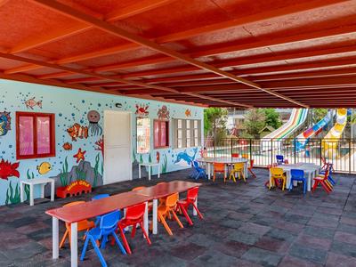 Überdachter Kinderspielbereich mit bunten Tischen, Stühlen und einer Wasserrutsche im Freien.