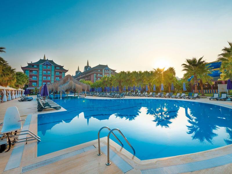 Resort pool with palm trees, loungers, and buildings under a clear blue sky at sunset.