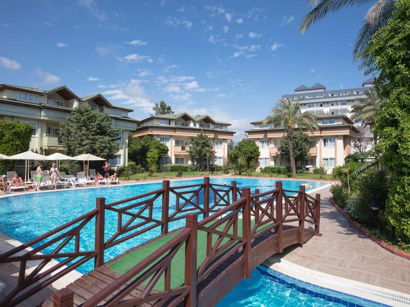 Resort pool area with wooden bridge, surrounded by lounge chairs and buildings under blue sky.