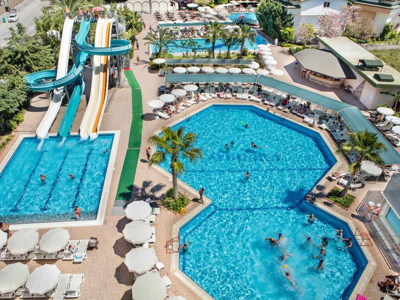 Resort pool area with slides, two large pools, palm trees, umbrellas, and lounge chairs.
