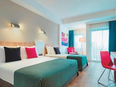 Hotelzimmer mit zwei Doppelbetten, weißen Wänden, bunten Kissen und Tisch mit rotem Stuhl.