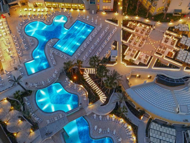 Přehled několika zářících modrých hotelových bazénů v noci s okolními budovami a cestami.