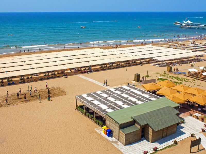 Pláž s slunečníky, plážovými bary a čistým modrým mořem za slunečného dne.