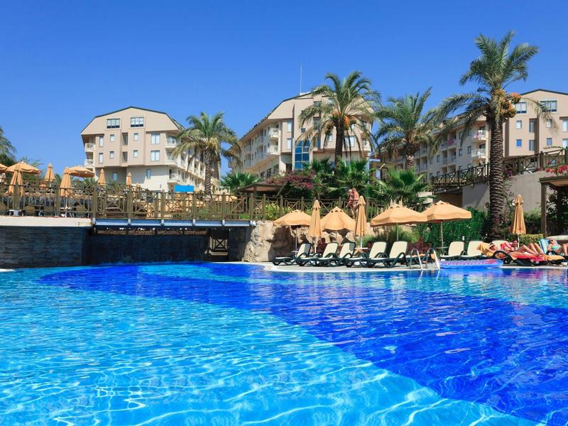 Velký hotelový bazén s lehátky a palmami pod jasnou modrou oblohou.