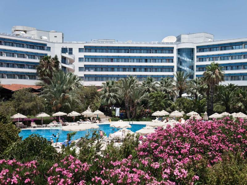 Complejo hotelero moderno con palmeras, piscina y arbustos rosas en flor al frente.