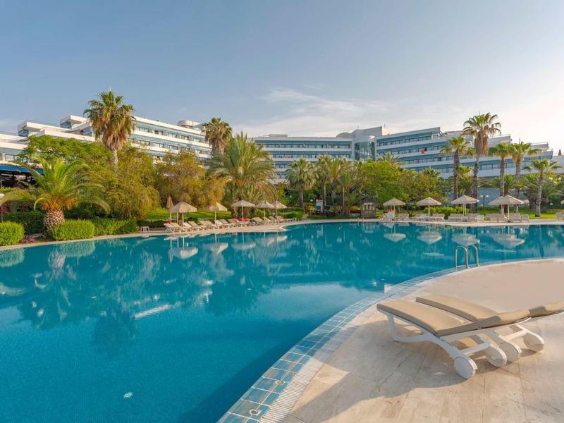 Gran piscina frente a un hotel moderno con palmeras y tumbonas bajo un cielo despejado.