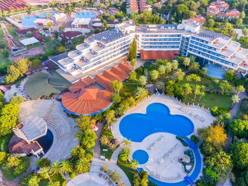 Vista aérea de un hotel con piscinas azules y rojas, rodeado de árboles y edificios.