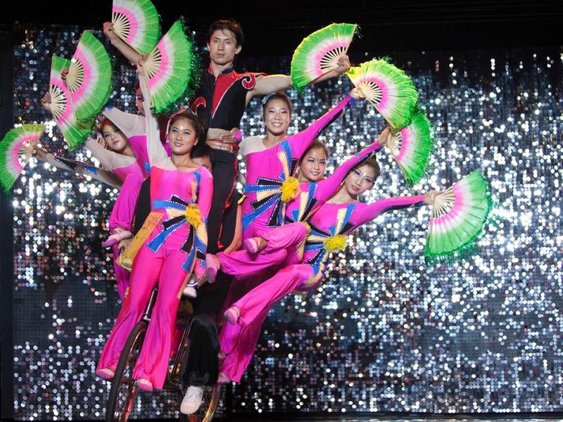 Grupo de bailarines en trajes coloridos con abanicos verdes en escenario brillante.