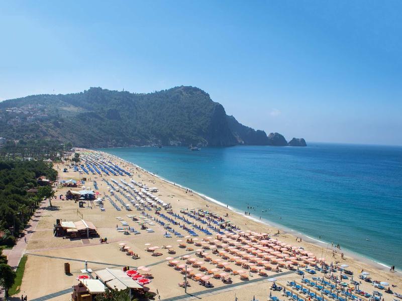 Lunga spiaggia sabbiosa con molti ombrelloni e mare turchese davanti a una collina boschiva.