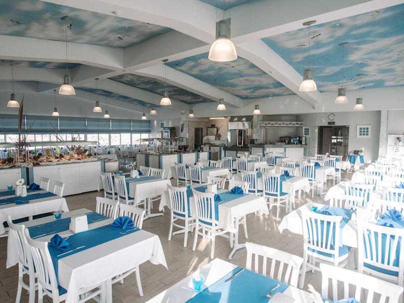 Ristorante luminoso e moderno con tavoli e sedie blu e bianchi sotto un soffitto dipinto con motivo cielo.